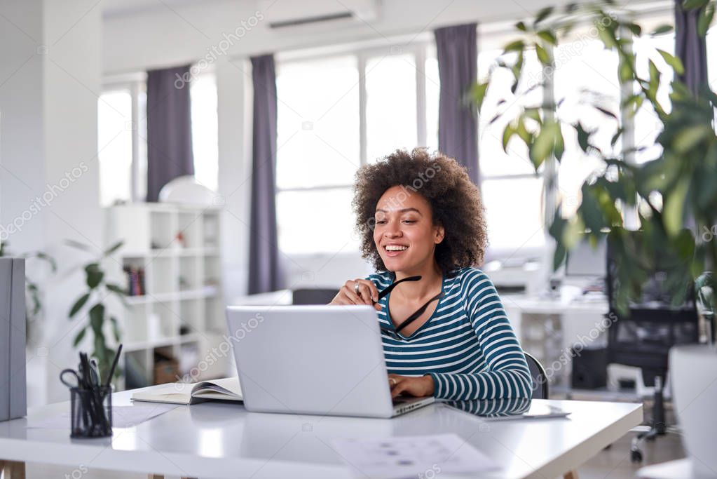 Smiling mixed race businesswoman holding eyeglasses and looking at laptop while sitting in modern office.