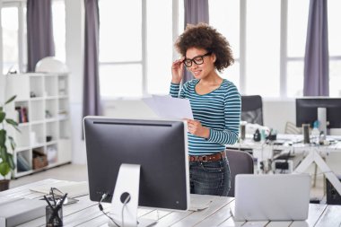 Güzel gülümseyen karışık ırk iş kadını ofiste rahat ayakta giyinmiş ve peperwork bakarak.