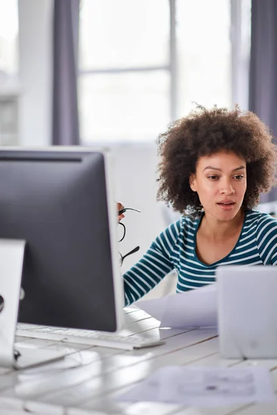 Attraktive Geschäftsfrau mit gemischter Rasse, lässig gekleidet, sitzt im Büro, hält Papiere in der Hand und blickt auf den Computermonitor. — Stockfoto