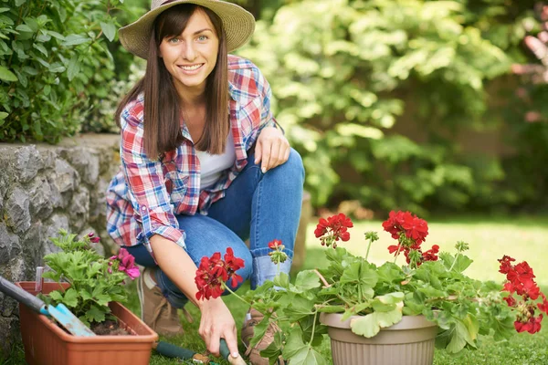 作業服を着た美しいコーカサスの女性がしゃがみ、裏庭で手熊手を使用しています。周りに女性は花の入った鉢. — ストック写真