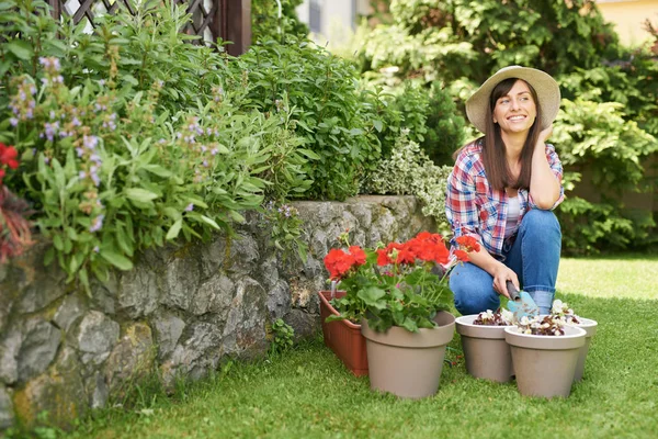 Morena encantadora em roupas de trabalho e com chapéu na cabeça agachado e segurando ferramenta de jardinagem. Na frente dela estão vasos com begônia e pelargonia . — Fotografia de Stock