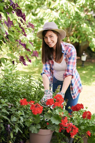 Linda morena branca sorridente em roupas de trabalho e com chapéu podando flores em seu quintal . — Fotografia de Stock