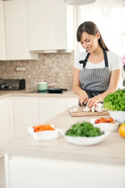 Όμορφο χαμόγελο αφιερωμένη Καυκάσιος μελαχρινή με ποδιά στέκεται στην κουζίνα και κόβοντας τα μανιτάρια. Στο τραπέζι είναι πολλά λαχανικά. Μαγείρεμα στο σπίτι ιδέα. — Φωτογραφία Αρχείου