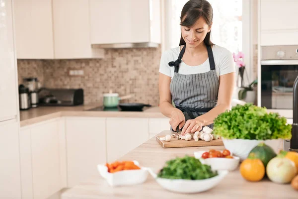 Linda morena caucasiana dedicada sorridente em avental de pé na cozinha e cortando cogumelos. Na mesa há muitos vegetais. Cozinhar em casa conceito . — Fotografia de Stock