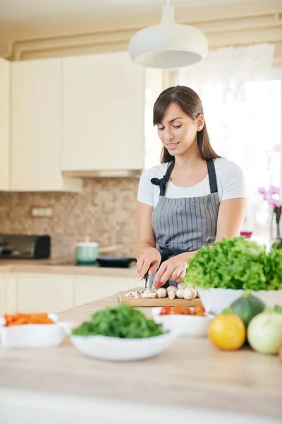 Mooie glimlachend toegewijde Kaukasische brunette in schort staand in de keuken en het hakken van paddestoelen. Op tafel zijn veel groenten. Koken bij Home concept. — Stockfoto