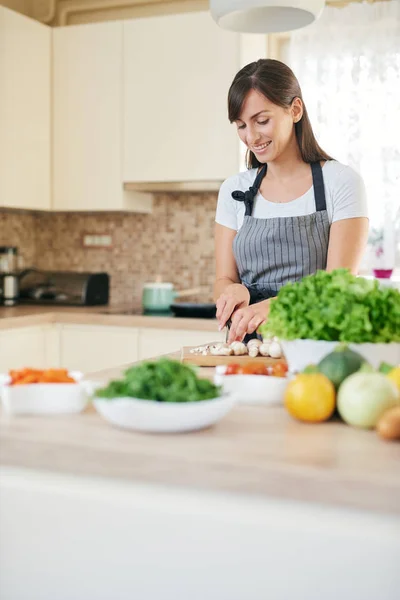 美丽的微笑专用的白种人布鲁内特在围裙站在厨房和切碎蘑菇。桌子上有很多蔬菜。在家烹饪概念. — 图库照片