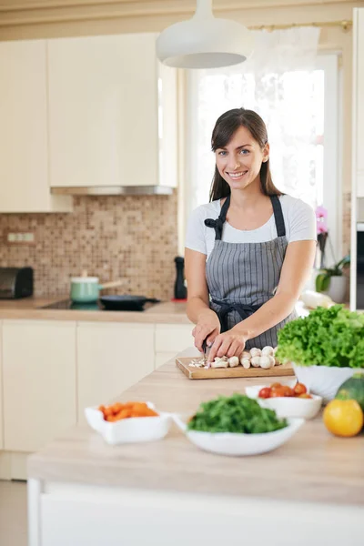 Linda morena caucasiana dedicada sorridente em avental de pé na cozinha e cortando cogumelos. Na mesa há muitos vegetais. Cozinhar em casa conceito . — Fotografia de Stock