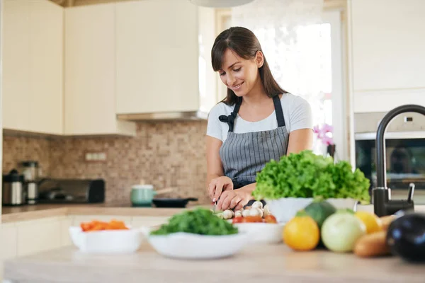 Linda morena caucasiana dedicada sorridente em avental de pé na cozinha e cortando cogumelos. Na mesa há muitos vegetais. Cozinhar em casa conceito . — Fotografia de Stock