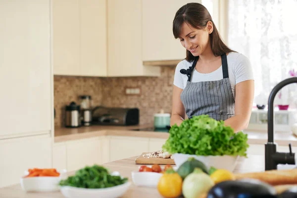 Linda morena caucasiana dedicada sorridente em avental de pé na cozinha e cortando cogumelos. Na mesa há muitos vegetais. Cozinhar em casa conceito . — Fotografia de Stock