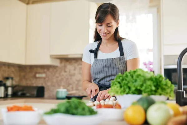 Linda morena caucasiana dedicada sorridente em avental de pé na cozinha e cortando cogumelos. Na mesa há muitos vegetais. Cozinhar em casa conceito . — Fotografia de Stock