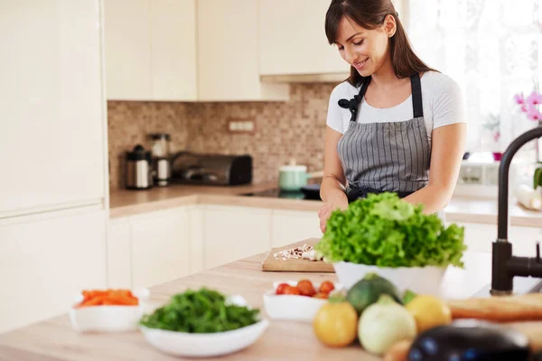 Linda morena caucasiana dedicada sorridente em avental de pé na cozinha e cortando cogumelos. Na mesa há muitos vegetais. Cozinhar em casa conceito . — Fotografia de Stock