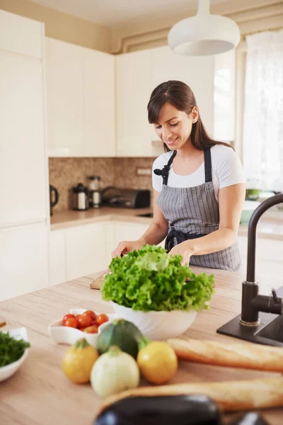 Mooie glimlachend toegewijde Kaukasische brunette in schort staand in de keuken en het hakken van paddestoelen. Op tafel zijn veel groenten. Koken bij Home concept. — Stockfoto