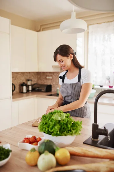 Linda morena caucasiana dedicada sorridente em avental de pé na cozinha e cortando cogumelos. Na mesa há muitos vegetais. Cozinhar em casa conceito . — Fotografia de Stock