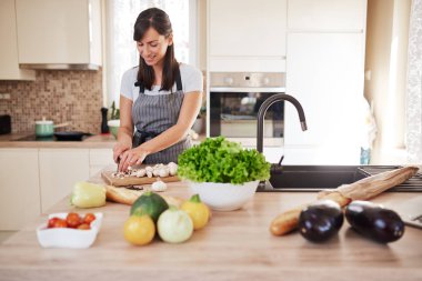 Güzel gülümseyen özel Kafkas esmer önlük mutfakta ayakta ve mantar doğrama. Masada bir sürü sebze var. Evde yemek pişirme konsepti.