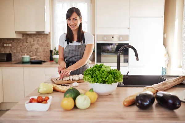 Vackra leende dedikerade kaukasiska brunett i förkläde stående i köket och hugga svamp. På bordet finns massor av grönsaker. Matlagning hemma Concept. — Stockfoto