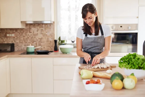 Όμορφο χαμόγελο αφιερωμένη Καυκάσιος μελαχρινή με ποδιά στέκεται στην κουζίνα και κόβοντας τα μανιτάρια. Στο τραπέζι είναι πολλά λαχανικά. Μαγείρεμα στο σπίτι ιδέα. — Φωτογραφία Αρχείου