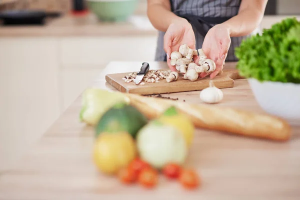 Mujer caucásica en delantal sosteniendo setas picadas mientras está de pie en la cocina. En la mesa de la cocina hay verduras y baguette . — Foto de Stock