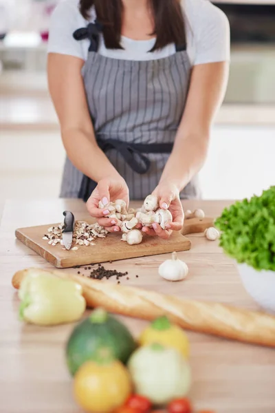 Kaukasische vrouw in schort houden gehakte paddestoelen terwijl staan in de keuken. Op keuken tafel zijn groenten en stokbrood. — Stockfoto