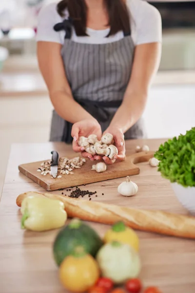 Kaukasische vrouw in schort houden gehakte paddestoelen terwijl staan in de keuken. Op keuken tafel zijn groenten en stokbrood. — Stockfoto