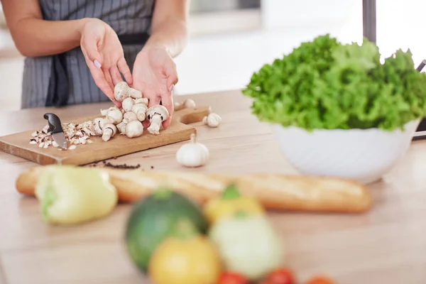 Toegewijde Kaukasische huisvrouw in schort staande in de keuken en het hakken van paddestoelen. Op tafel zijn veel groenten. Koken bij Home concept. — Stockfoto