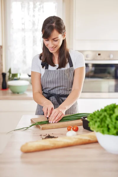 Leende kaukasiska kvinnlig i förkläde hugga purjolök i köket och förbereder middag. Beredning av inhemsk matkoncept. — Stockfoto