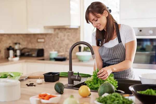 Mulher caucasiana atraente alegre no avental que lava a salada na cozinha. Jantar em casa conceito . — Fotografia de Stock