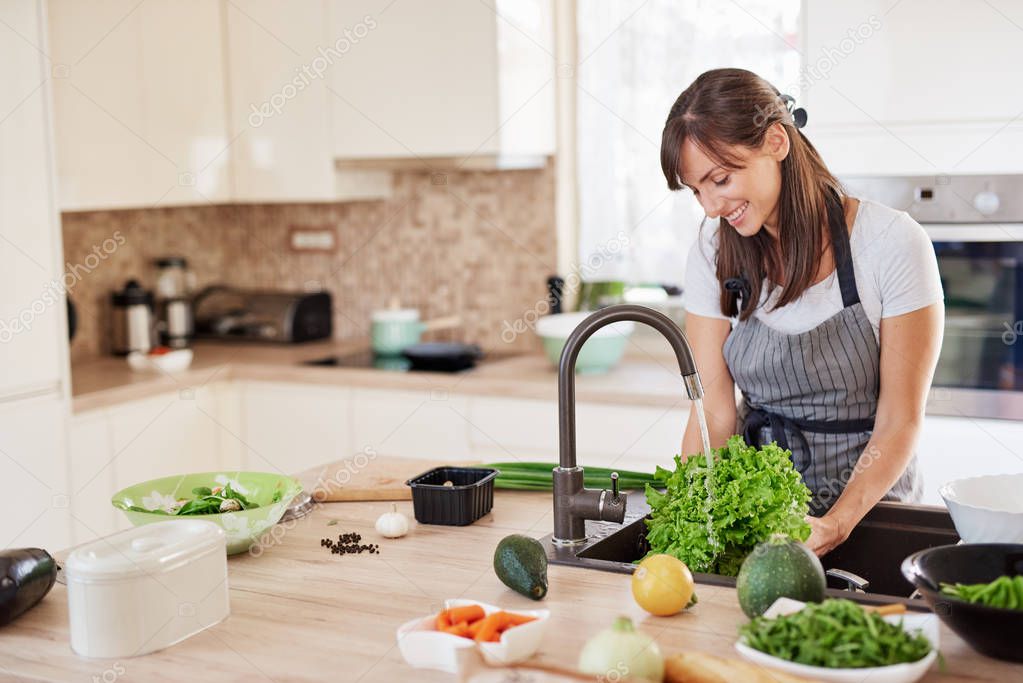 Cheerful attractive Caucasian woman in apron washing salad in kitchen. Dinner at home concept.