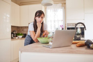 Güzel Kafkas esmer önlük mutfak tezgahına yaslanmış ve dizüstü bakarak ve akşam yemeği için tarifi aşağıdaki. Elinde havuç tutan kadın.