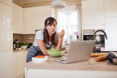 Mutfak tezgahına yaslanmış, dizüstü bilgisayara bakan, yeni bir tarif arayan ve havuç yiyen önlükteki muhteşem Kafkas esmeri. Masada bir sürü sebze var..