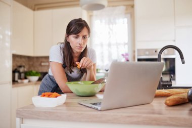 Güzel Kafkas esmer önlük mutfak tezgahına yaslanmış ve dizüstü bakarak ve akşam yemeği için tarifi aşağıdaki. Elinde havuç tutan kadın.