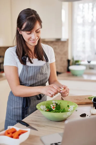 Linda morena caucasiana em avental fazendo salada de legumes para o jantar ou almoço e seguindo a receita no laptop. Interior da cozinha doméstica . — Fotografia de Stock