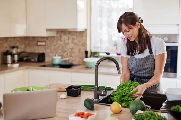 Mulher caucasiana atraente alegre no avental que lava a salada na cozinha. Jantar em casa conceito . — Fotografia de Stock