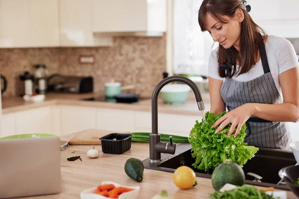 Χαρούμενη ελκυστική καυκάσια γυναίκα με ποδιά πλύσιμο σαλάτα στην κουζίνα. Δείπνο στο σπίτι ιδέα. — Φωτογραφία Αρχείου