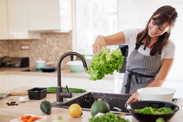 Vrolijke aantrekkelijke Kaukasische vrouw in schort wassen salade in de keuken. Dinner at Home concept. — Stockfoto