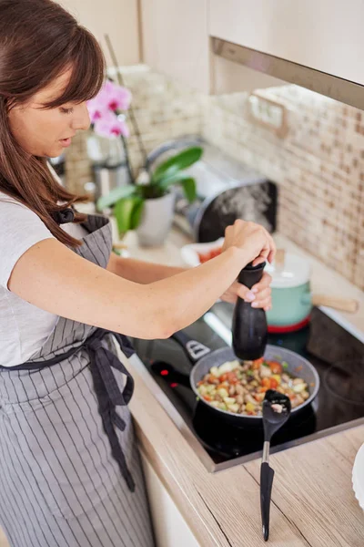 Charming Caucasain morena em avental de pé ao lado do fogão na cozinha e adicionando pimenta em legumes cozidos jantar . — Fotografia de Stock