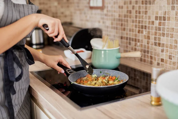 Charming Caucasain brunette in apron standing next to stove in kitchen and cooking healthy dinner. — Stock Photo, Image