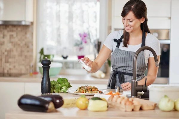 Morena branca bonito em avental de pé na cozinha e adicionando sal em massa deliciosa fresca. Preparação do conceito de alimento italiano . — Fotografia de Stock