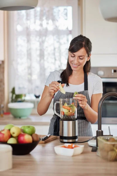 Vackra kaukasiska leende kvinna i förkläde sätta grönsaker och frukter i mixer medan du står i köket. — Stockfoto