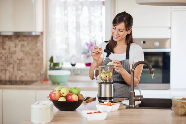 Bella donna caucasica sorridente in grembiule mettere verdure e frutta in frullatore mentre in piedi in cucina . — Foto Stock