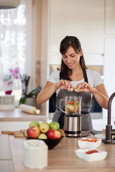 Vackra kaukasiska leende kvinna i förkläde sätta grönsaker och frukter i mixer medan du står i köket. — Stockfoto