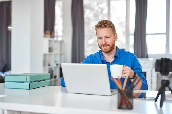 Unga dedikerade kaukasiska manliga anställd sitter i kontoret, dricka kaffe och använda laptop. Starta affärsidé. — Stockfoto