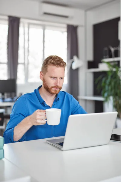 Joven empleado masculino caucásico dedicado sentado en la oficina, bebiendo café y utilizando el ordenador portátil. Concepto de creación de empresas . — Foto de Stock