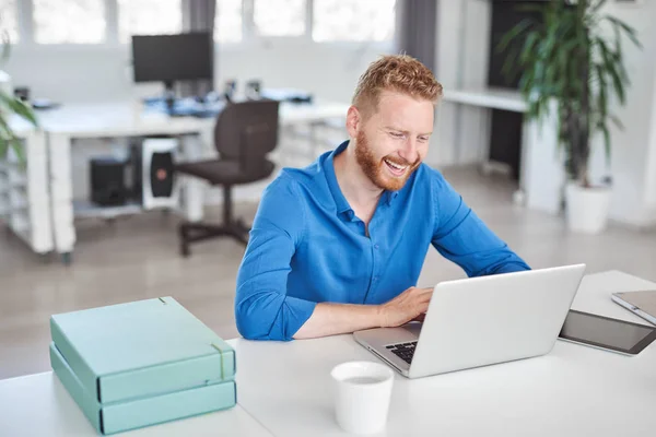 Junge lächelnde gutaussehende kaukasische männliche Angestellte, die im Büro sitzt und Laptop benutzt. Existenzgründungskonzept. — Stockfoto