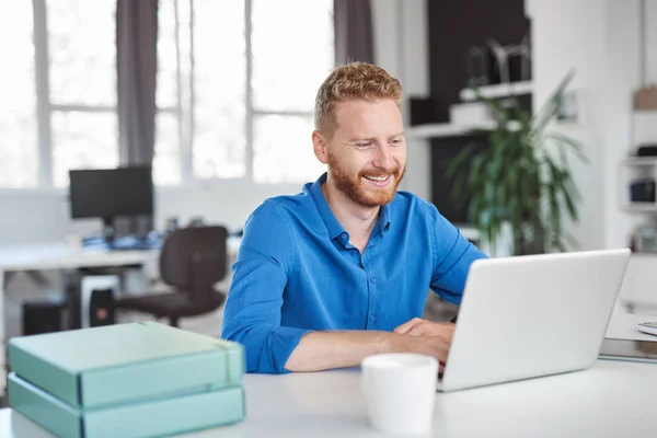 Giovane sorridente bel dipendente maschio caucasico seduto in ufficio e utilizzando il computer portatile. Concetto di start up . — Foto Stock