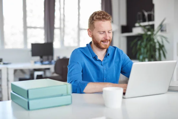 Unga leende vacker kaukasiska manliga anställd sitter på kontoret och använder laptop. Starta affärsidé. — Stockfoto