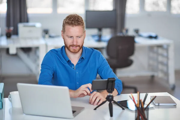 Jonge blanke medewerker met een videogesprek via een smartphone terwijl hij in kantoor zit. Op de Bureau is een laptop. Bedrijfsconcept opstarten. — Stockfoto
