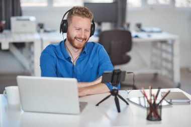 Genç Kafkas çalışanı ofiste otururken akıllı telefon üzerinden görüntülü görüşme yapıyor. Baş kulaklık ve masada dizüstü bilgisayar ve kahve.