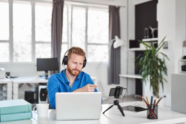 Genç Kafkas çalışanı ofiste otururken akıllı telefon üzerinden görüntülü görüşme yapıyor. Baş kulaklık ve masada dizüstü bilgisayar ve kahve.