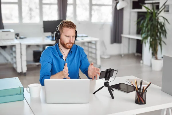 Joven empleado caucásico que tiene videollamada por teléfono inteligente mientras está sentado en la oficina. En la cabeza hay auriculares y en el escritorio es portátil y café . — Foto de Stock