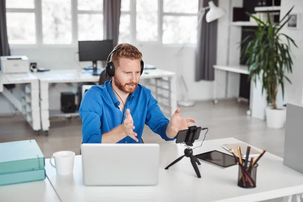 Jonge blanke werknemer met video-oproep via smartphone terwijl zittend in kantoor. Op het hoofd zijn hoofdtelefoons en op Bureau is laptop en koffie. — Stockfoto